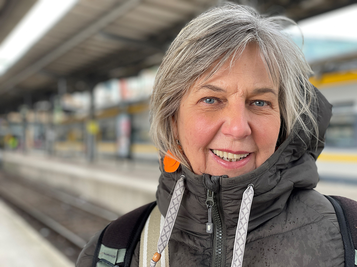 Maria Sigroth står i förgrunden i en grå täckjacka, i bakgrunden syns centralstationen med tåg och järnvägsspår.